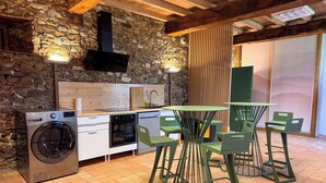 Eine Küche mit Steinwand, Holzbalken, einer Waschmaschine, einem Herd und einem Essbereich mit grünen Stühlen und einem Tisch.