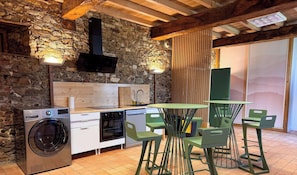 Une cuisine avec un mur en pierre, des poutres en bois, un lave-linge, une cuisinière et un coin repas avec des chaises vertes et une table.