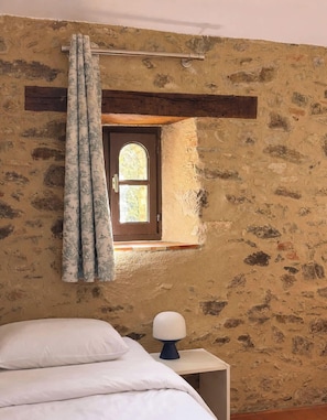 A room with a stone wall, a window with a wooden frame, a bed with white bedding, and a bedside table with a lamp.