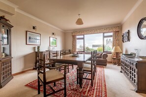 A spacious dining area with a large wooden table, multiple chairs, and a red patterned rug. There are framed pictures on the walls, a window with curtains, and a chandelier. - Melon Park | Elie & Earlsferry (Elie)
