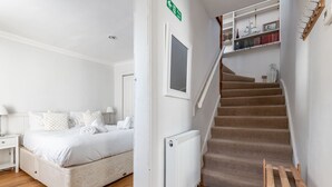 Een slaapkamer met een bed, nachtkastje, radiator, houten trap en een boekenplank.