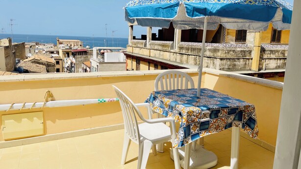 A balcony with a table and chairs, a patterned tablecloth, and a blue umbrella. - "La Tonnara" is a studio apartment with a sea view terrace, it is located in the central area (Castellammare del Golfo)