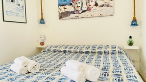 A neatly made bed with a blue and white patterned bedspread, a framed painting of two figures on a rocky shore, and a small shelf with a green bottle. - "La Tonnara" is a studio apartment with a sea view terrace, it is located in the central area (Castellammare del Golfo)