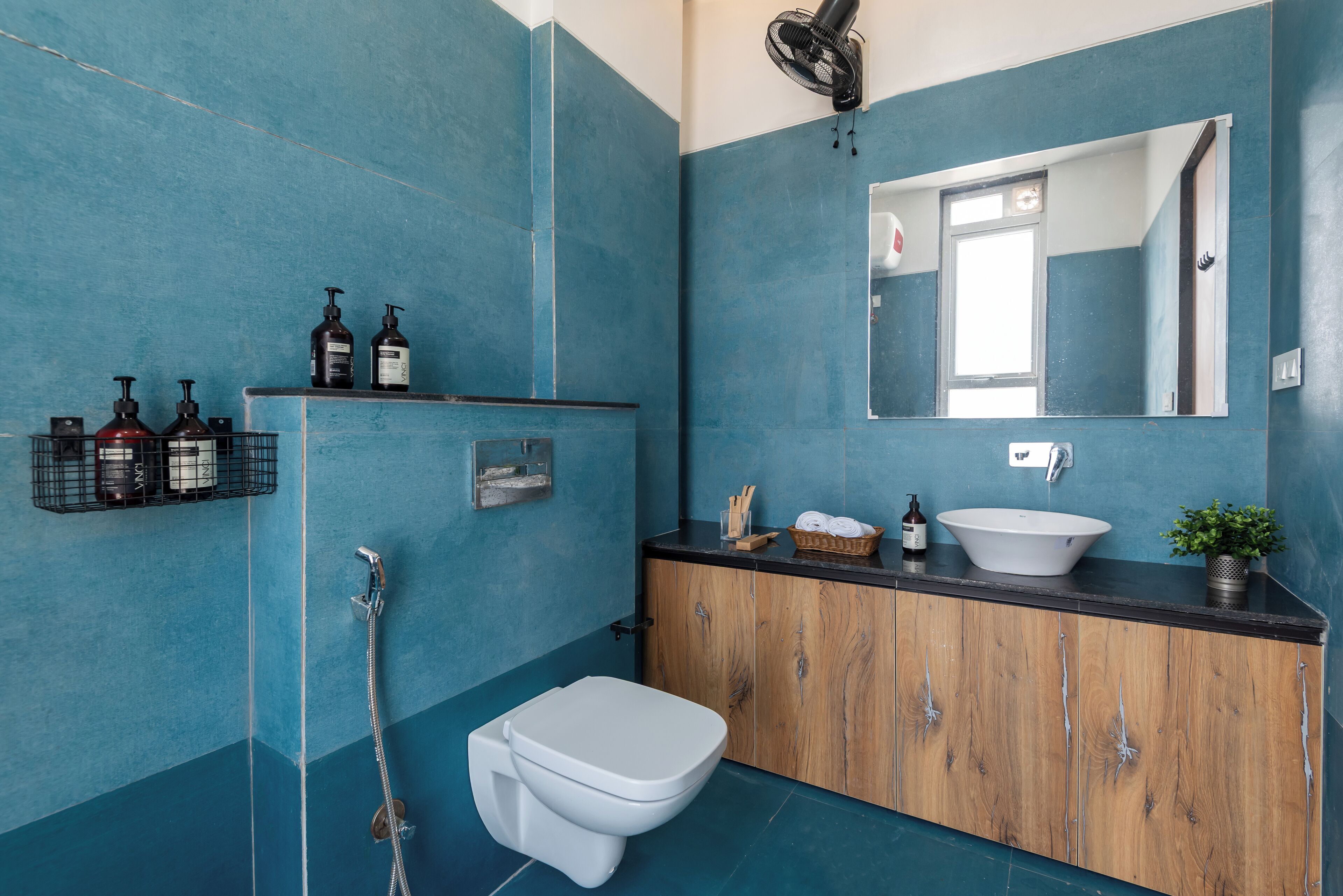Un baño con pared azul de azulejos, un inodoro blanco, un tocador de madera con lavabo y un espejo.