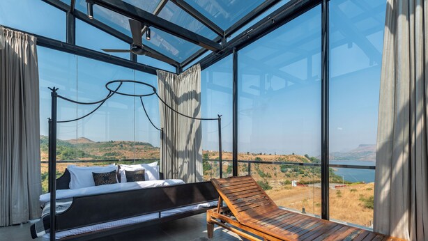 A modern bedroom with a bed, a wooden lounge chair, and a large glass window offering a view of a hilly landscape. - ELIVAAS White House 4BHK Villa With Pool (Igatpuri)
