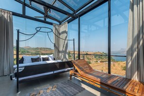 A modern bedroom with a bed, a wooden lounge chair, and a large glass window offering a view of a hilly landscape. - ELIVAAS White House 4BHK Villa With Pool (Igatpuri)