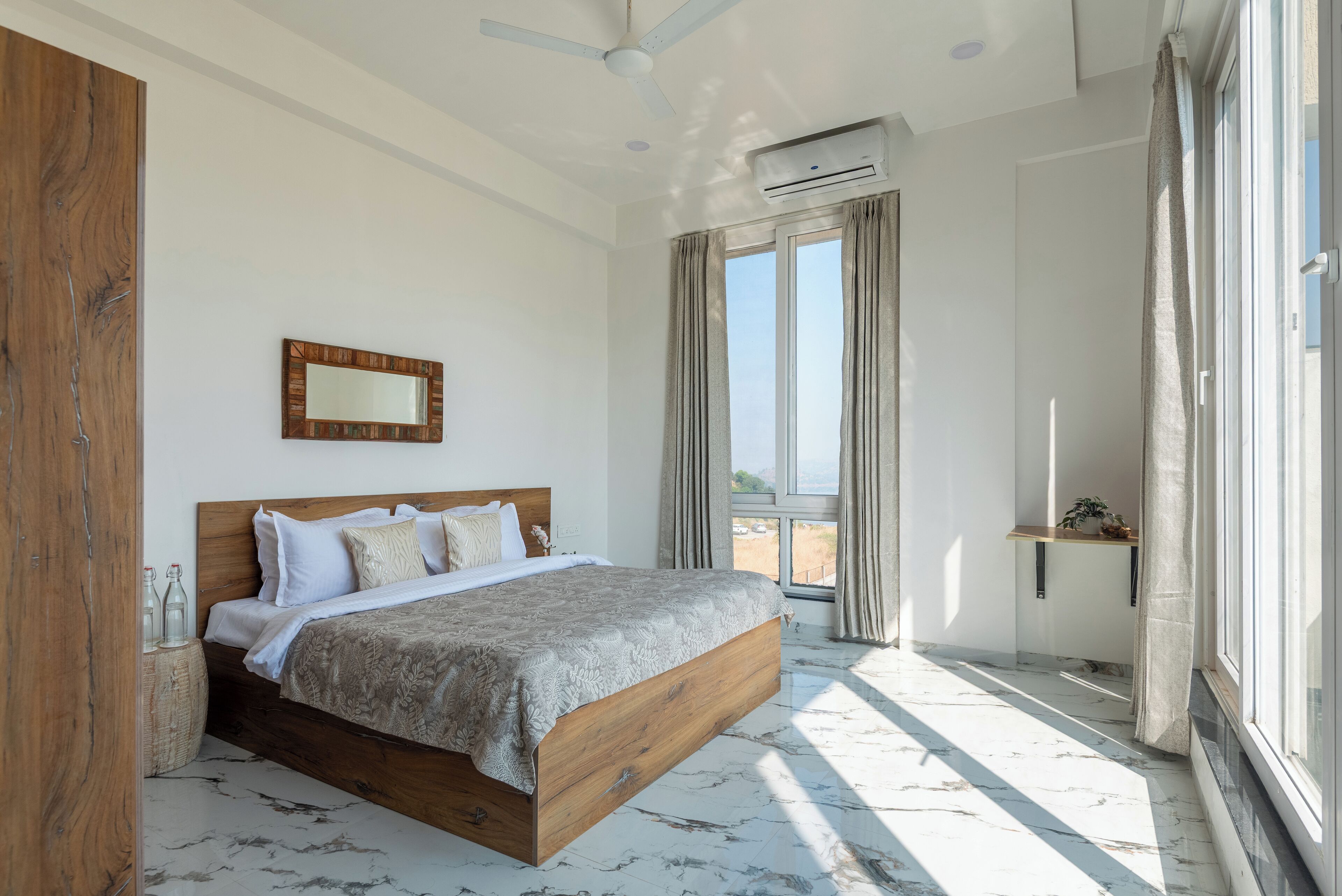 A bedroom with a large bed, a ceiling fan, an air conditioning unit, a window with curtains, and a small table with a plant.