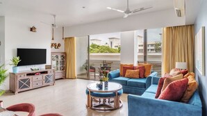 A spacious living room with a flat-screen TV, a blue sofa, a coffee table, and a dining area with red chairs. - Vita by Dia hotels And Homes  (Nerul)