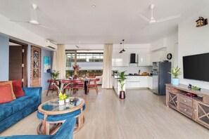A spacious living area with a blue sofa, a glass coffee table, and a dining area with a red table and chairs. A kitchen with white cabinets and a black refrigerator is visible in the background. - Vita by Dia hotels And Homes  (Nerul)