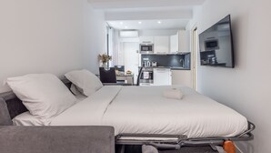 A compact hotel room with a bed, a desk, a chair, a small kitchenette, and a TV. - 2 PIECES MODERN FELIX FAURE A1B232 (Cannes)