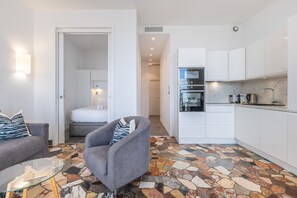 A compact living space with a sofa, armchair, and a glass coffee table. A bed is visible in the adjacent room, and a kitchen with white cabinets and a marble countertop is in the background. - 2 BEDROOMS—BALCONY—OCEAN VIEW—FELIX FAURE A2B231 (Cannes)