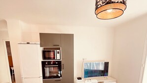 A modern kitchen with a built-in oven, microwave, and refrigerator. There is a dining table with chairs and a TV on a stand. - 🏆 T4 NEUF CENTRE-VILLE - 3 CHAMBRES - LIONS DE CANNES 2026 5 mn des Plages (Cannes)