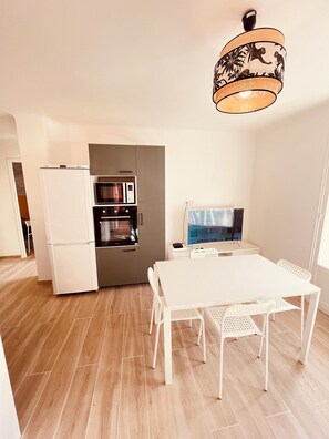 A modern kitchen with a built-in oven, microwave, and refrigerator. There is a dining table with chairs and a TV on a stand. - 🏆 T4 NEUF CENTRE-VILLE - 3 CHAMBRES - LIONS DE CANNES 2026 5 mn des Plages (Cannes)