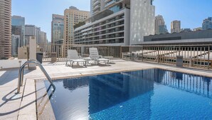Piscina en la azotea con una escalera metálica, sillas reclinables blancas y vistas a un paisaje urbano.