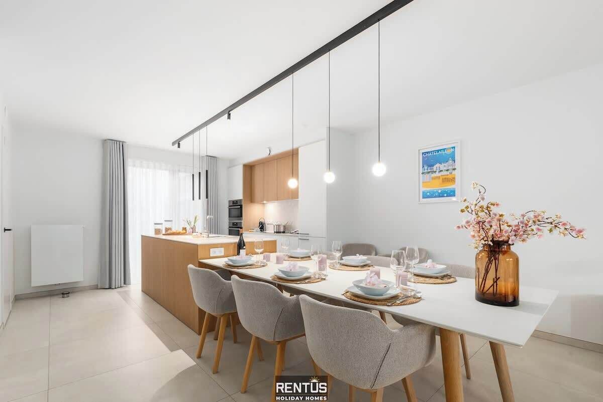 A modern dining area with a white table set for four, grey chairs, and a vase with flowers on the right.