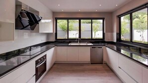 A modern kitchen with a black countertop, white cabinets, a built-in oven, and a large window providing a view of the outdoors. - วิลลาพร้อมสระว่ายน้ำ 4 ห้องนอน 4 ห้องน้ำ พร้อมสิ่งอำนวยความสะดวก (Tambon San Phak Wan)