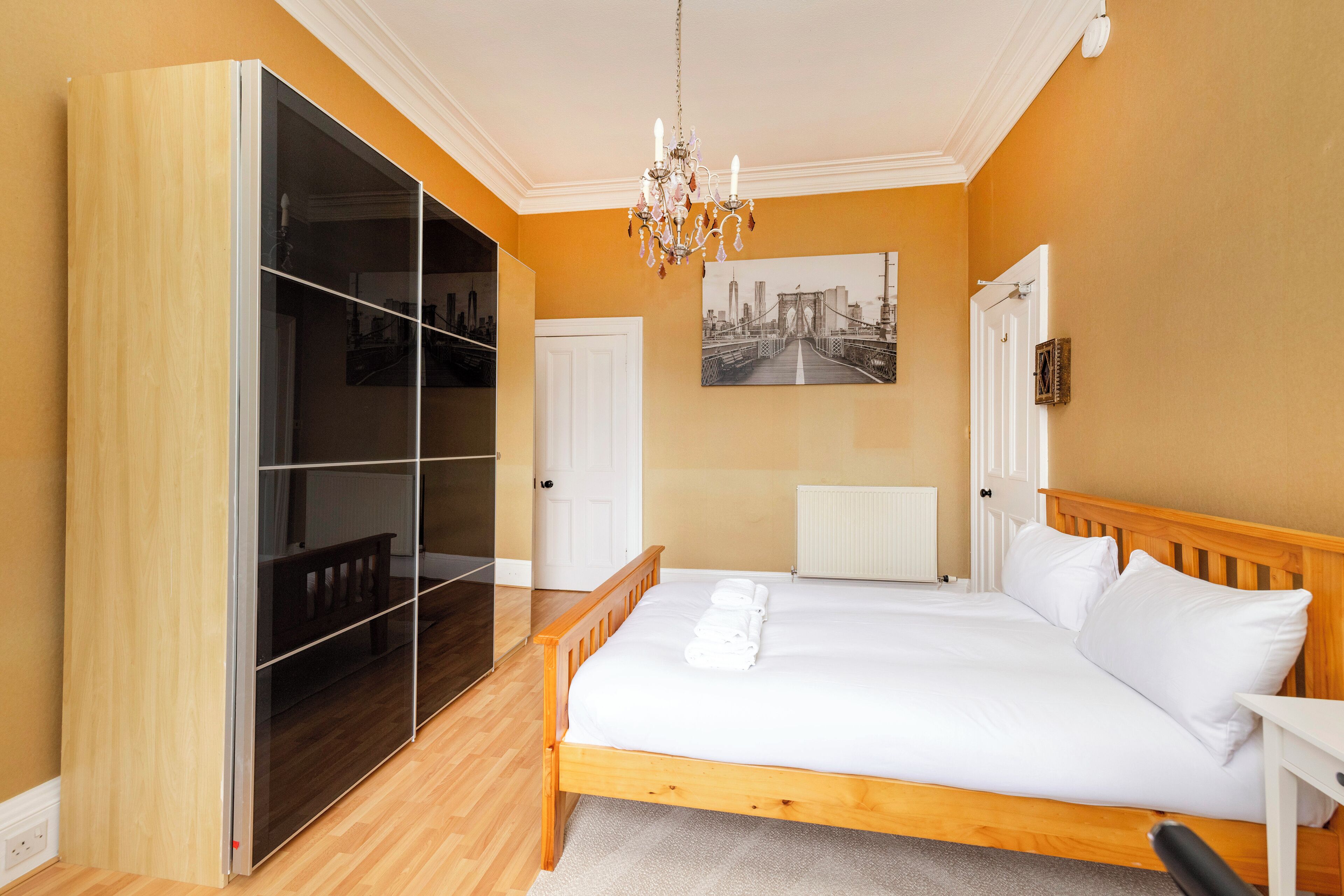 Een slaapkamer met een houten bed, een grote garderobe, een kroonluchter en een stadsgezichtschilderij aan de muur.