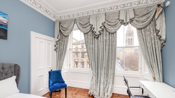 A hotel room with a bed, a blue armchair, a desk, and a view of a cityscape through large windows with curtains. - Stylish Southside Flat Near Old Town (Edinburgh)