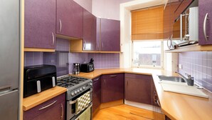 A kitchen with purple cabinets, a gas stove, a toaster, and a window with blinds. - Stylish Southside Flat Near Old Town (Edinburgh)