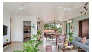 A spacious living room with a sofa, ottomans, a coffee table, and a dining area with chairs and a table. There are ceiling fans, a TV, and a view of the outdoors. - Viora by Dia hotels And Homes  (Nerul)