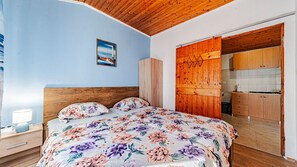 A bedroom with a wooden ceiling, a bed with floral bedding, a nightstand with a lamp, and a view of a kitchen through an open door.