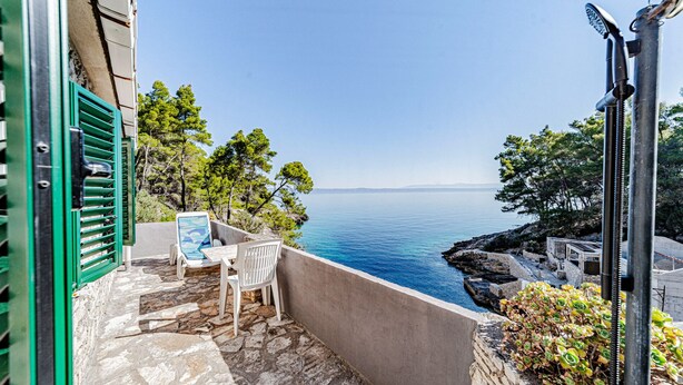Un balcon donnant sur la mer, une chaise, une table et une fenêtre à volets verts.