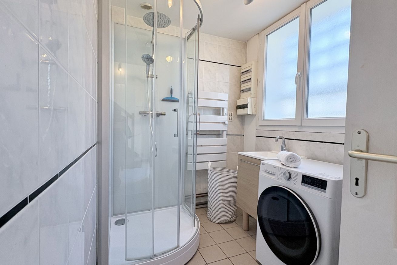 Une salle de bain moderne dotée d’une douche à parois de verre, d’un lave-linge encastré et d’une fenêtre laissant entrer la lumière naturelle.