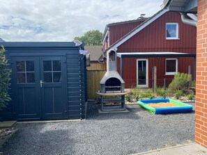 Ein Garten mit einem Ziegelhaus, einem schwarzen Geräteschuppen und einem Pizzaofen aus Stein.