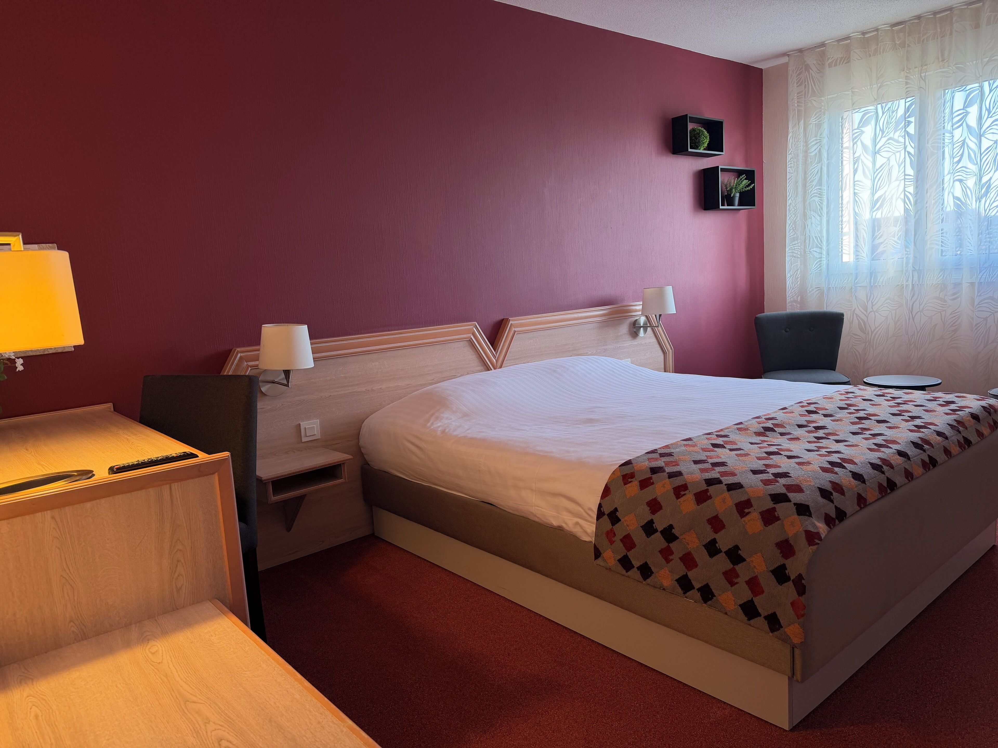 A hotel room with a bed, bedside tables, a chair, a small desk, and a window with curtains.
