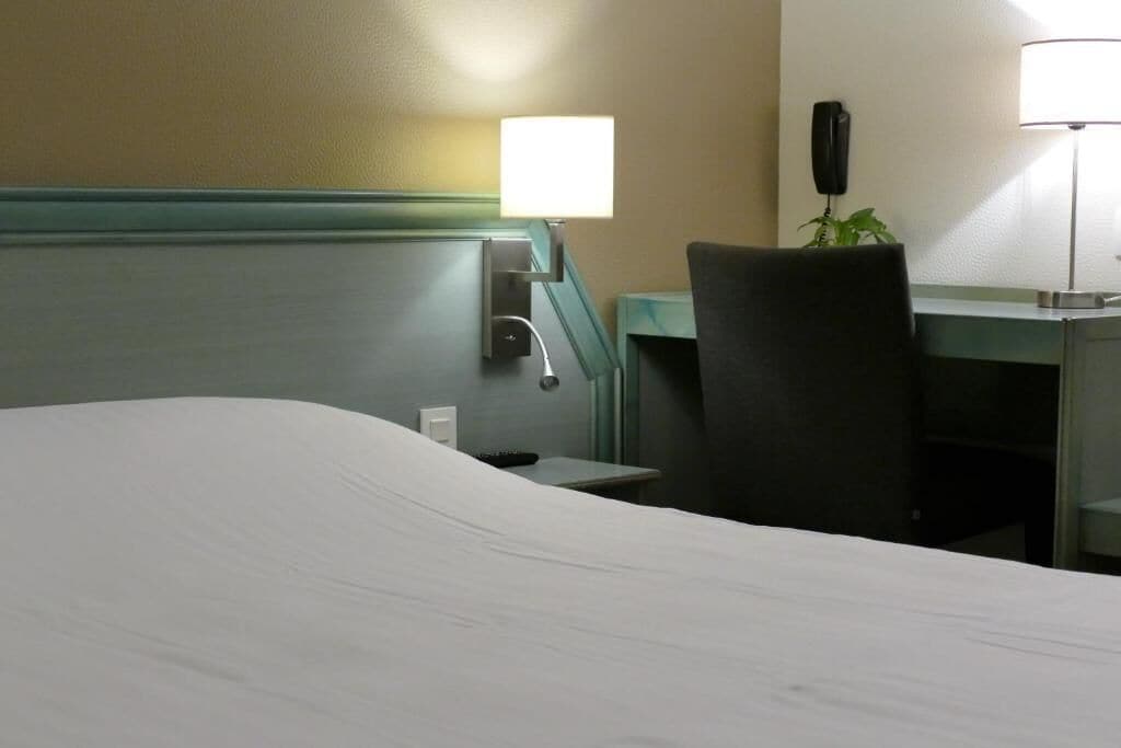 A hotel room with a bed, a desk, a chair, a lamp, and a phone on the wall.