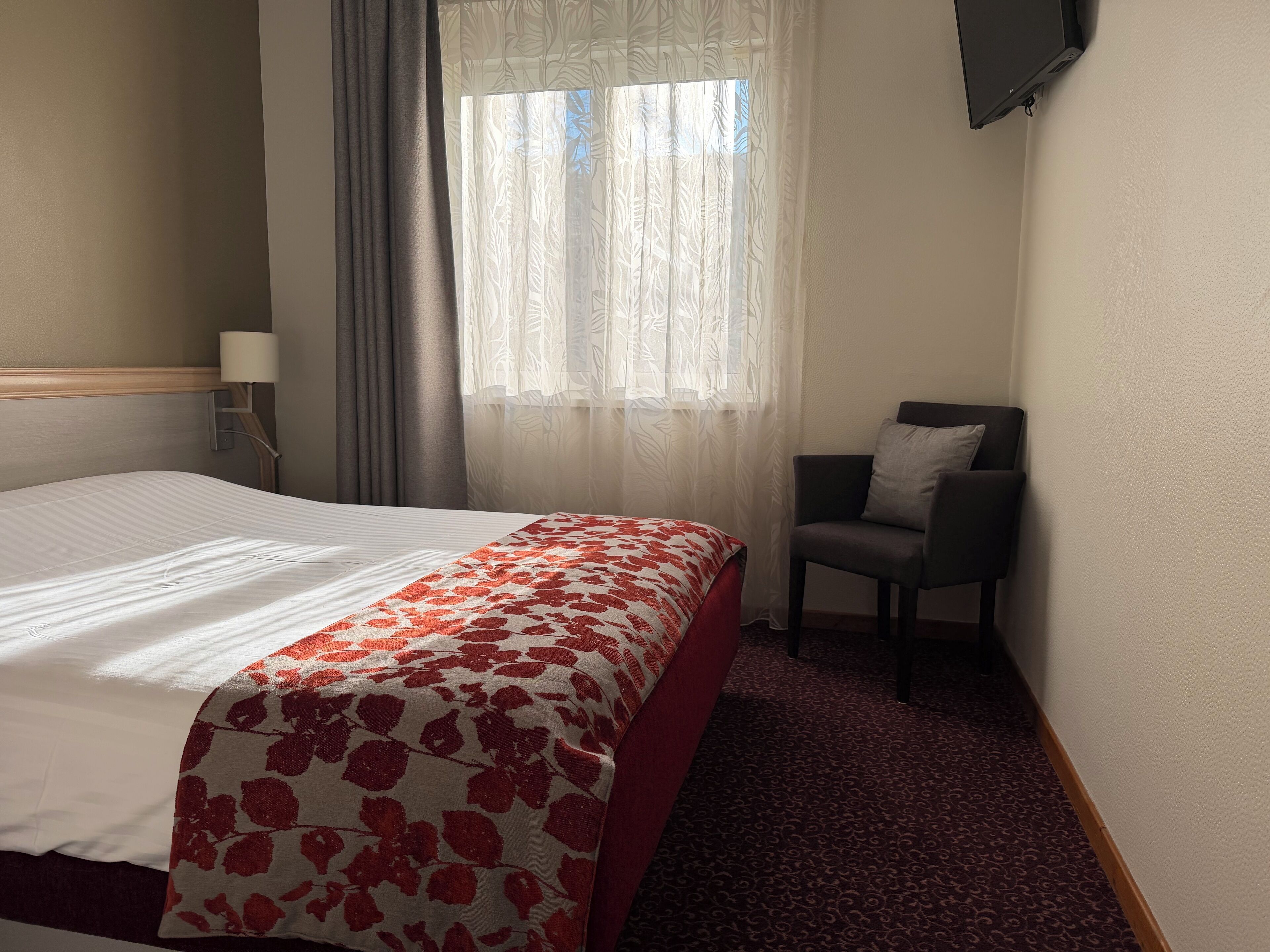 A hotel room with a bed, a chair, a TV, a window with curtains, and a lamp.