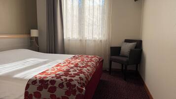 A hotel room with a bed, a chair, a TV, a window with curtains, and a lamp.