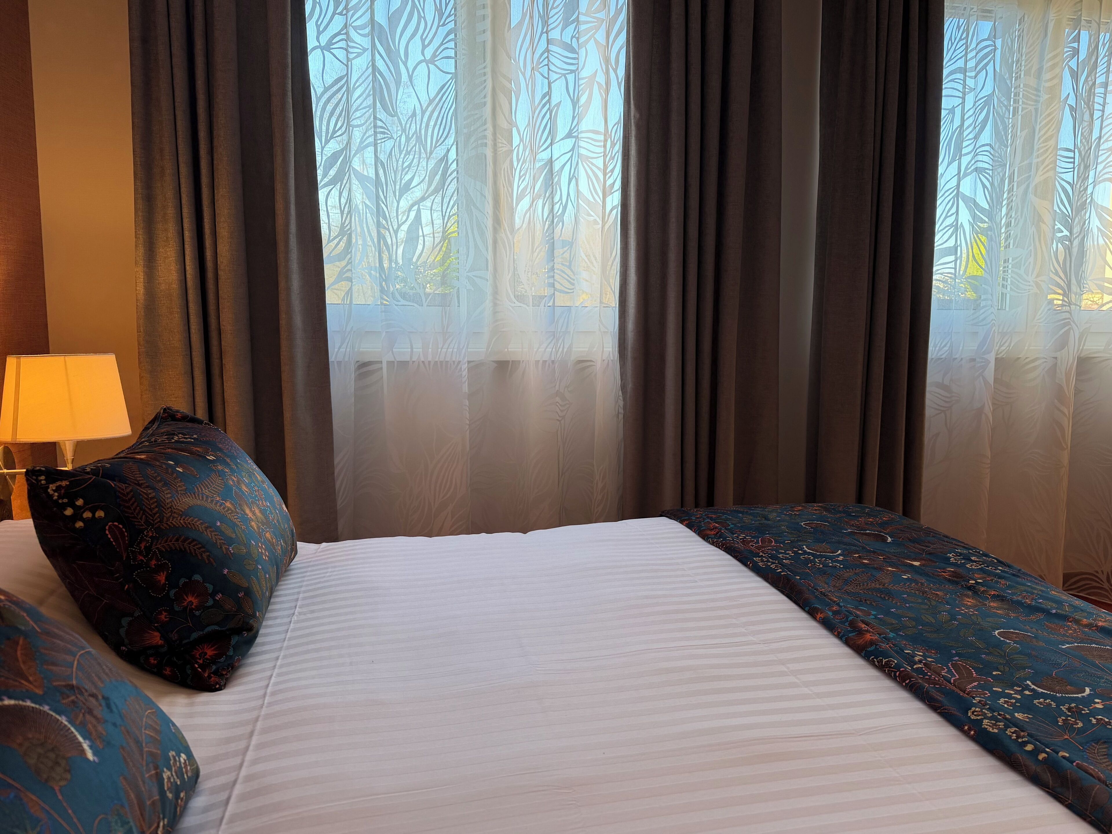 A hotel room with a bed, a lamp, and sheer curtains with a leaf pattern.