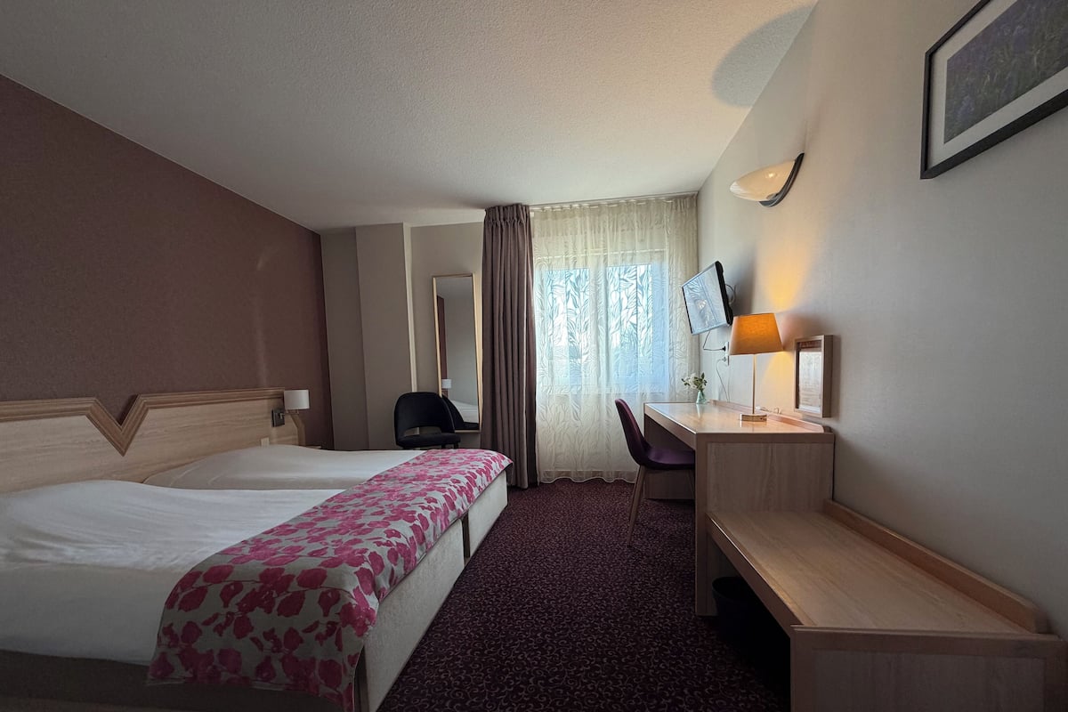 A hotel room with two beds, a desk, a chair, a desk lamp, a TV, and a window with curtains.