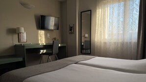 Une chambre d’hôtel avec deux lits, un bureau, une chaise, une télévision, un miroir et une fenêtre avec des rideaux légers.