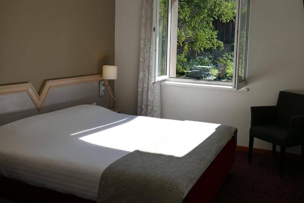 Une chambre d’hôtel avec un lit, une chaise, une fenêtre donnant sur un espace verdoyant et une lampe sur la table de chevet.