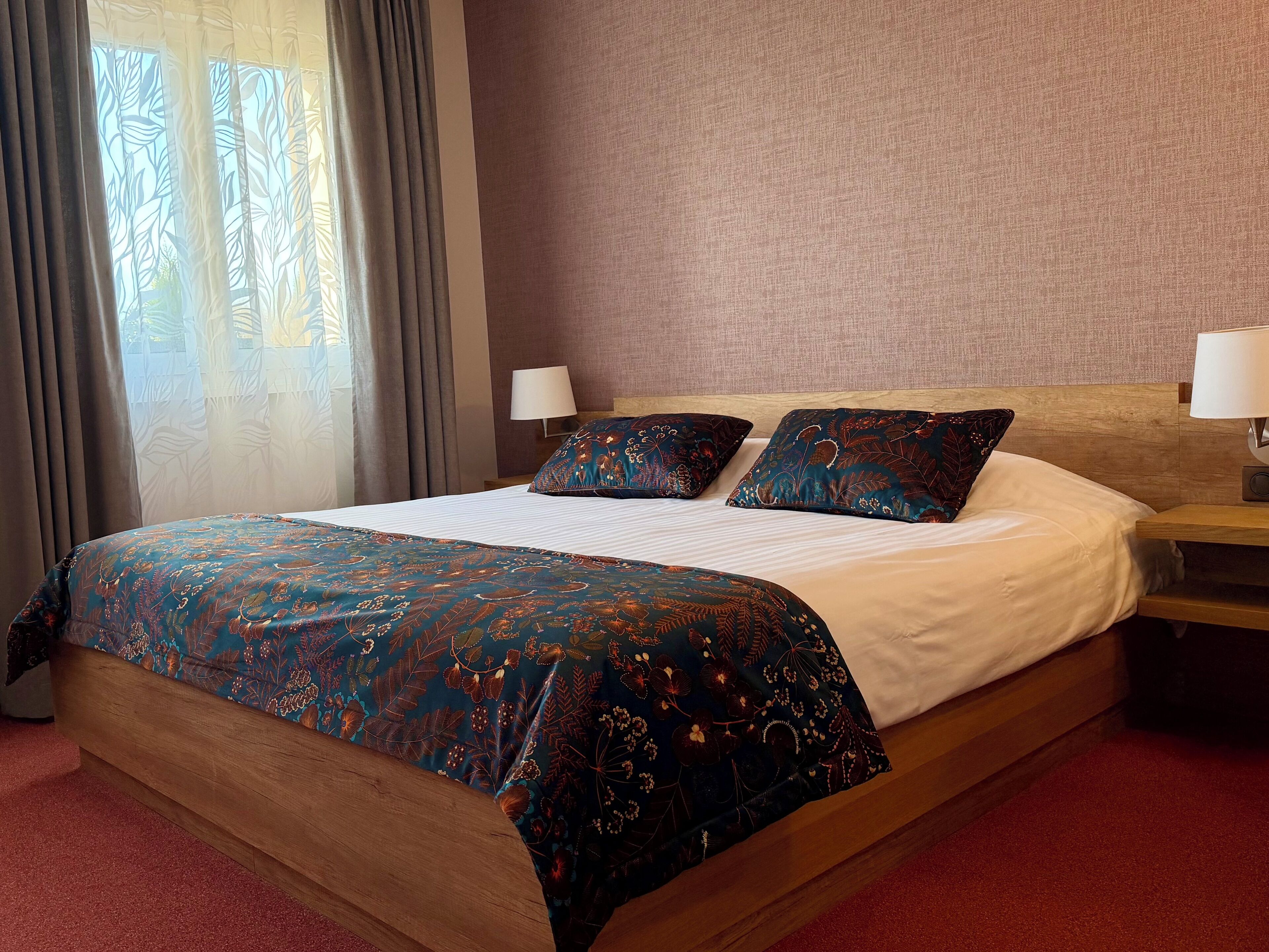 Habitación de hotel con una cama grande, dos almohadas, una mesita de noche, una lámpara y un ventanal con cortinas.