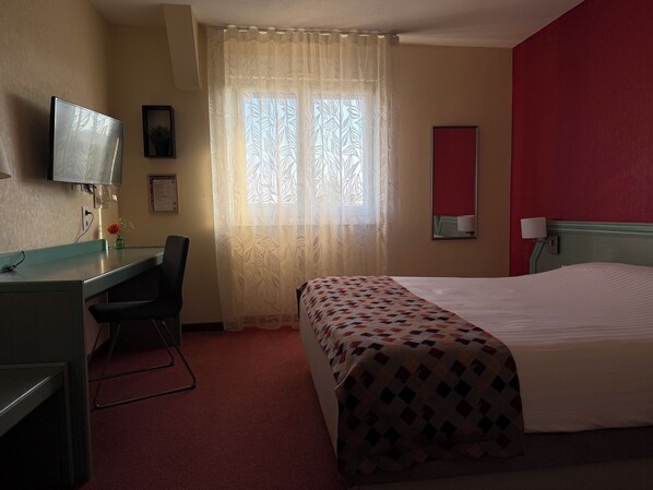 A hotel room with a bed, a desk, a chair, a television, a window with a curtain, and a red wall.