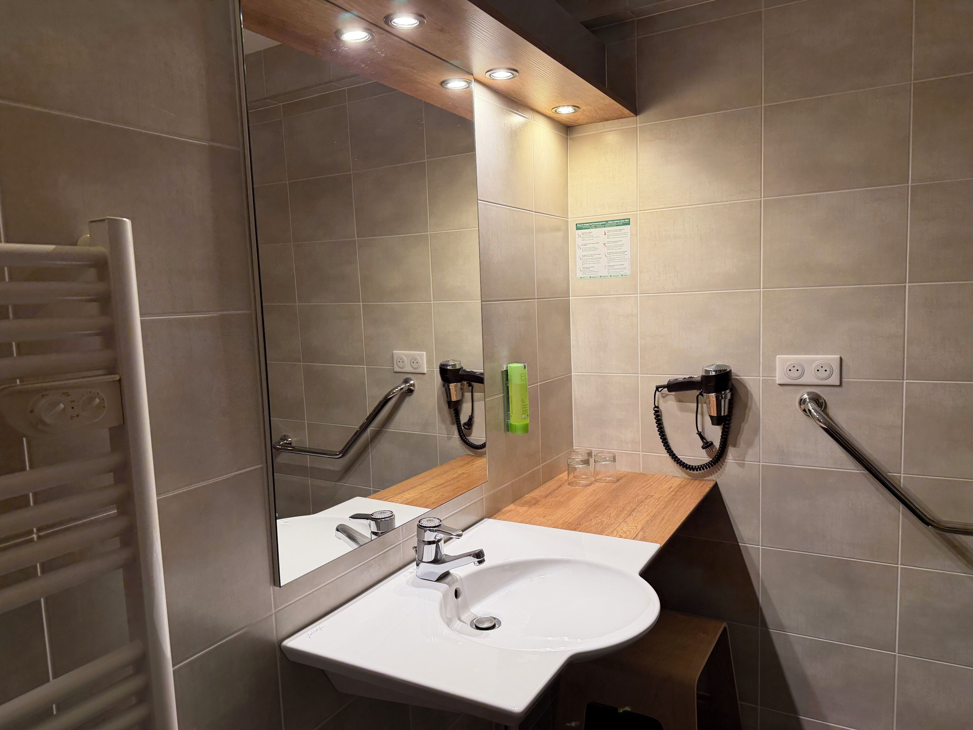 A bathroom with a white sink, mirror, and towel rack. There are two hair dryers mounted on the wall, and a wooden countertop.