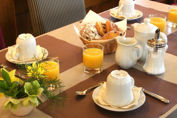 Une table de petit-déjeuner avec un panier de viennoiseries, un pichet, une théière, un pot à sucre et un verre de jus d’orange.