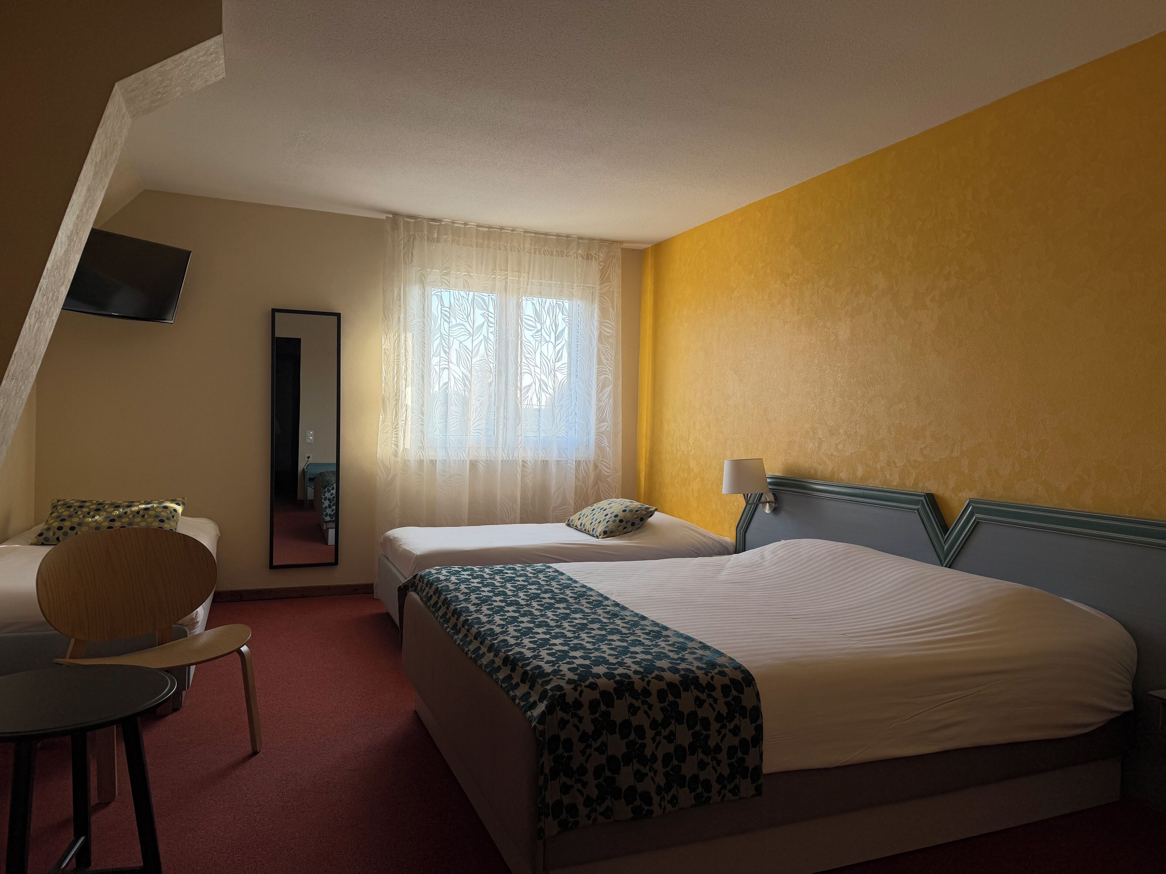 A hotel room with a bed, a chair, a desk, a mirror, and a window with curtains.