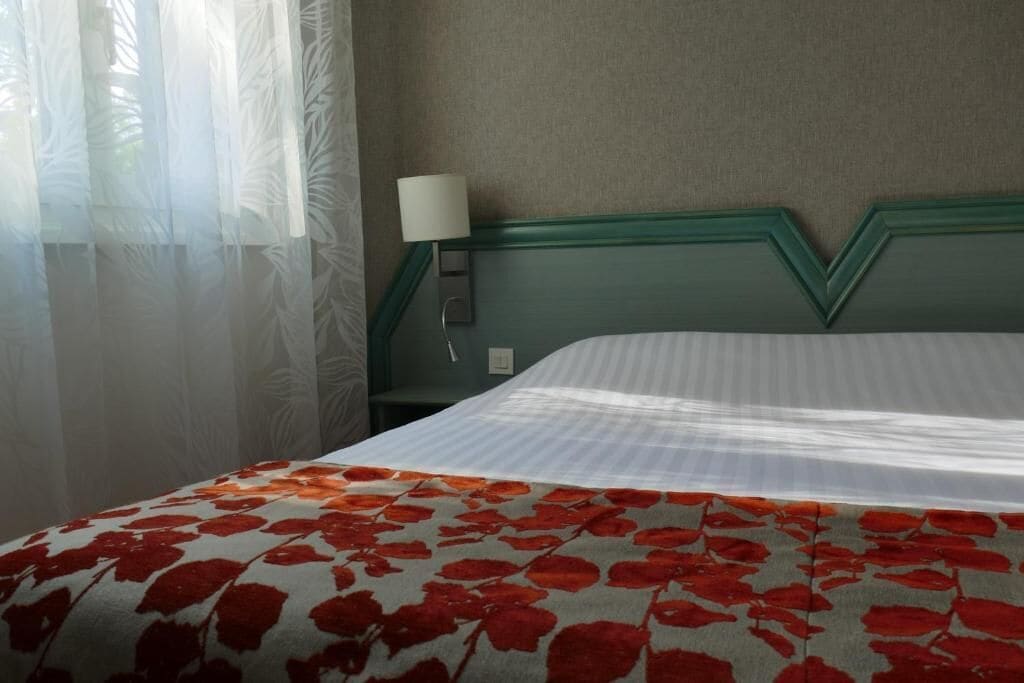Een hotelkamer met een bed, een nachtkastje met een lamp en dunne gordijnen.