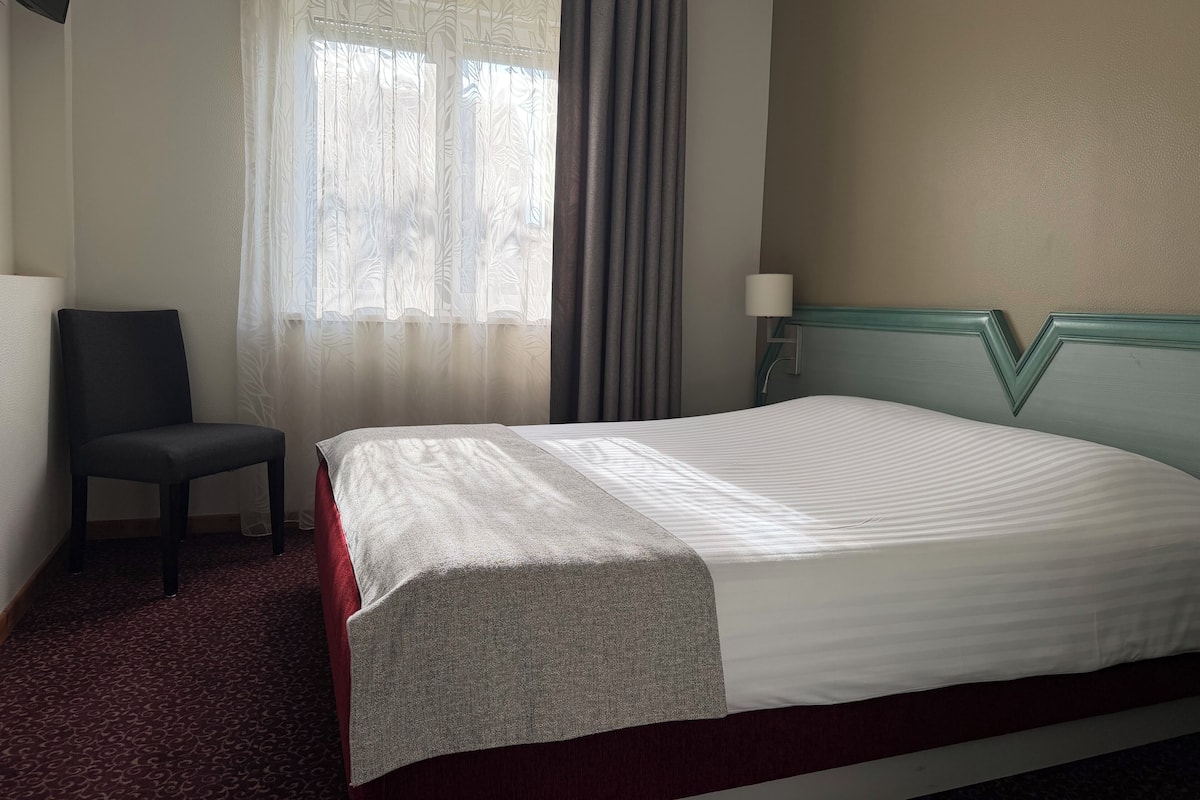 A hotel room with a bed, a chair, a window with curtains, and a lamp.
