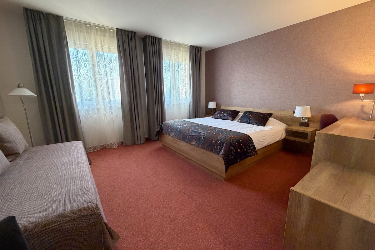 A hotel room with a bed, a desk, a chair, a lamp, and a window with curtains.