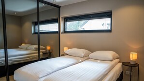 A modern hotel room with a large bed, a bedside table with a lamp, a window with blinds, and a mirror reflecting the room.