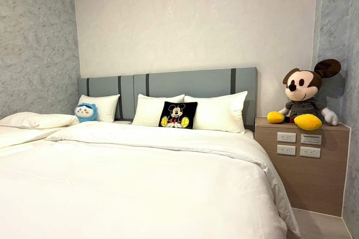 Une chambre d’hôtel avec un lit, des oreillers, une peluche à l’effigie de Mickey Mouse et une table de chevet avec une lampe.