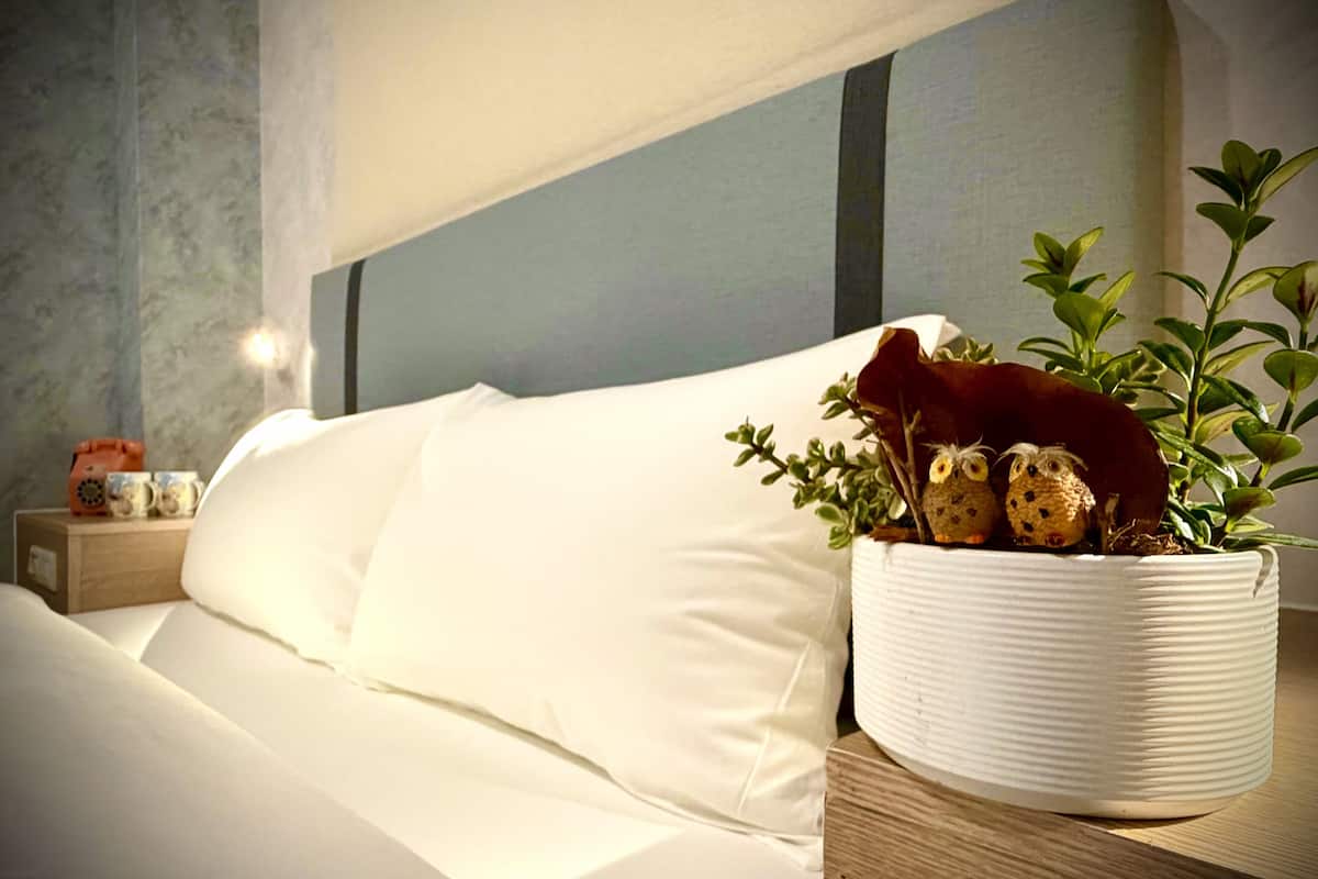 Un lit bien fait, avec du linge de lit blanc, une table de chevet sur laquelle se trouve un pot blanc contenant une plante, et un téléphone d’époque.