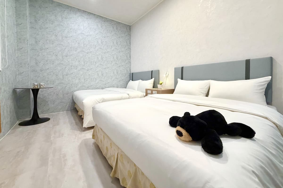 Une chambre d’hôtel avec deux lits, une petite table, un ours en peluche sur l’un des lits et une table de chevet avec une lampe.