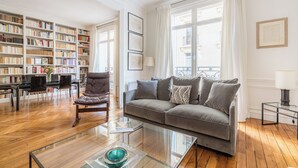 Sala de estar com sofá, mesa de centro de vidro, estante e área de jantar com cadeiras e mesa.