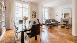 Uma sala de estar espaçosa com piso em madeira em padrão herringbone, uma mesa de jantar de vidro com cadeiras, um sofá e uma lareira.